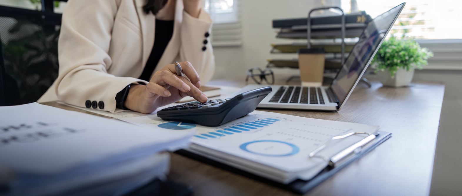 An accountant uses a calculator and laptop to reconcile figures at a bright desk.