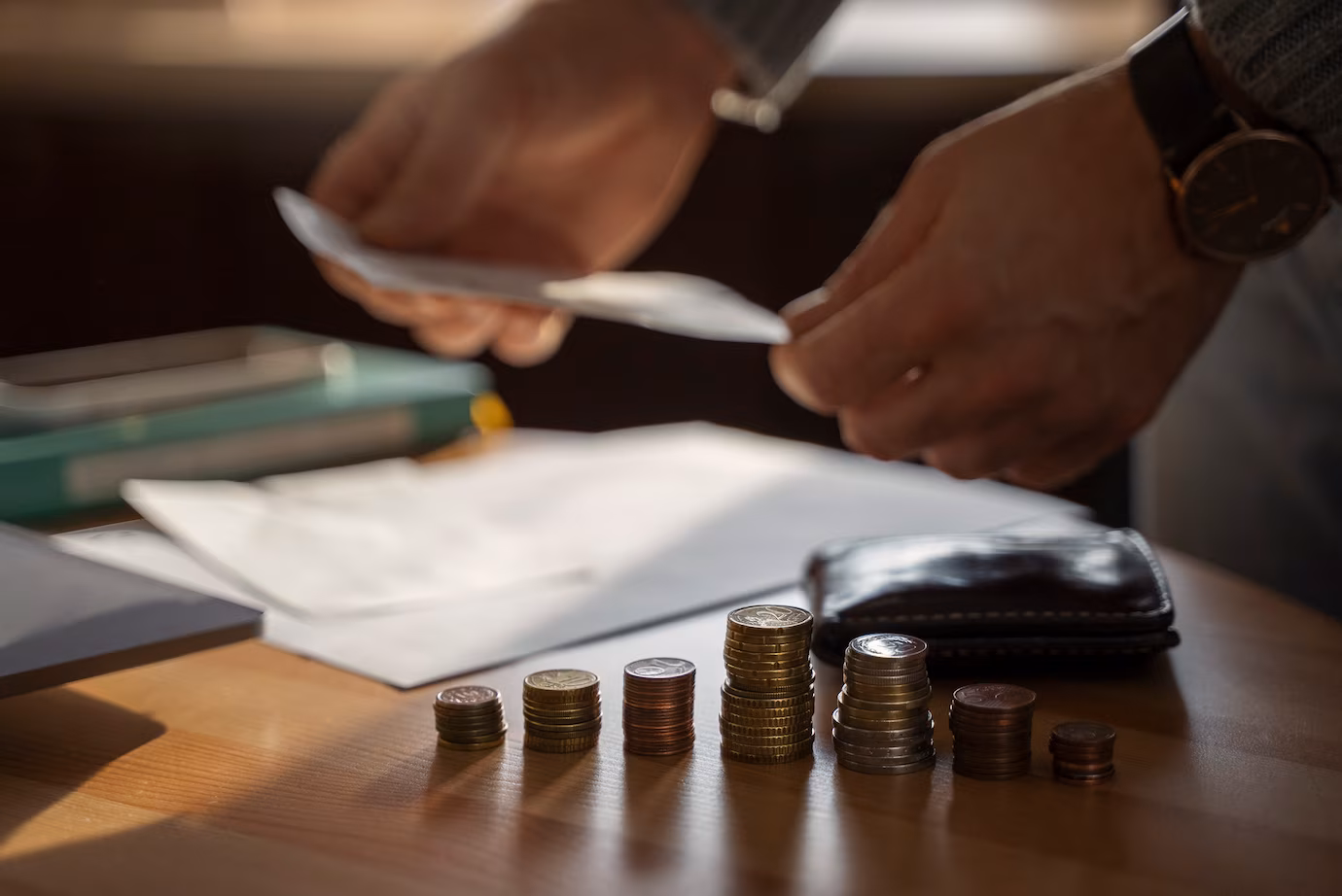Hands counting notes beside stacks of coins on a desk.