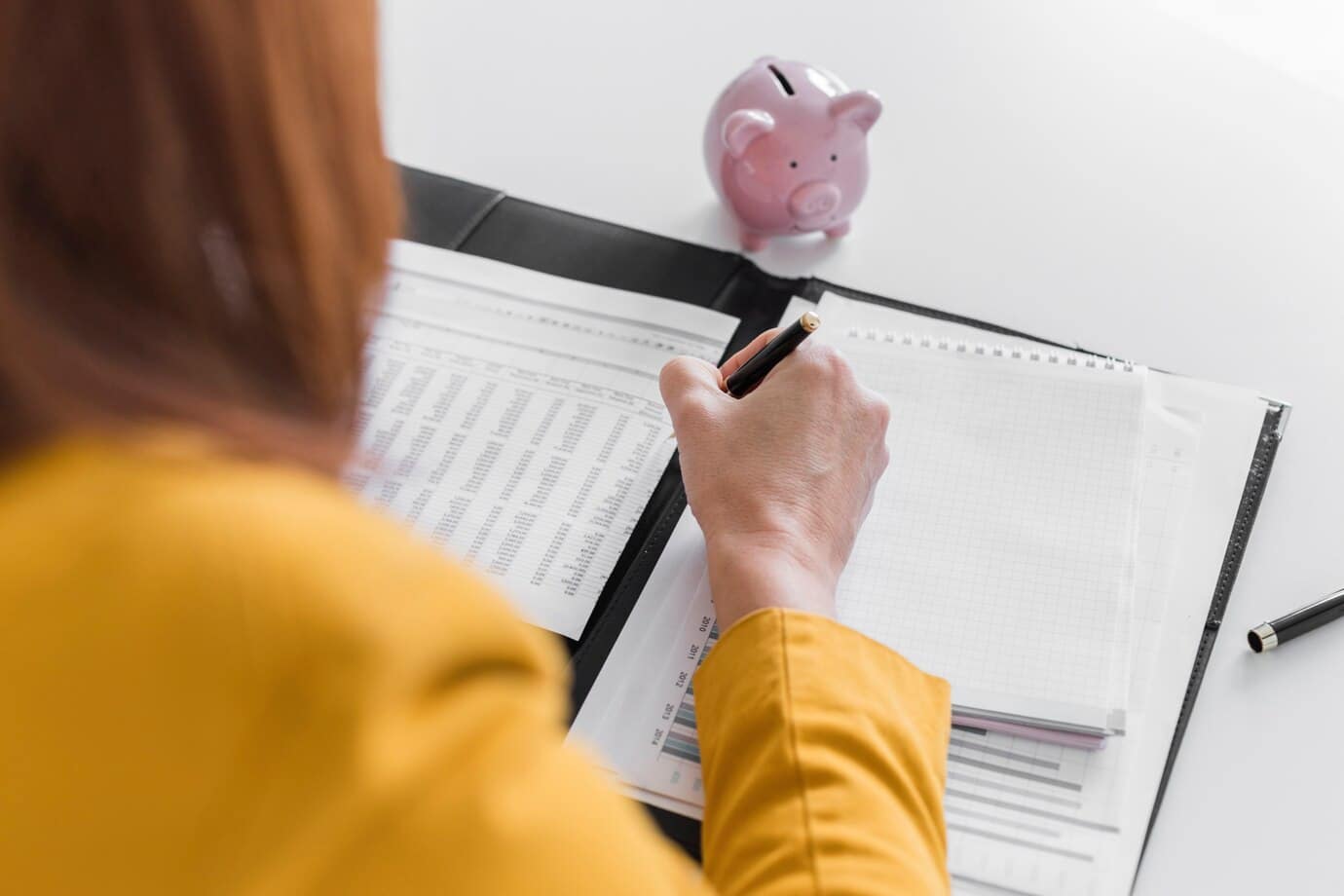 A person writing on a checklist beside a piggy bank.