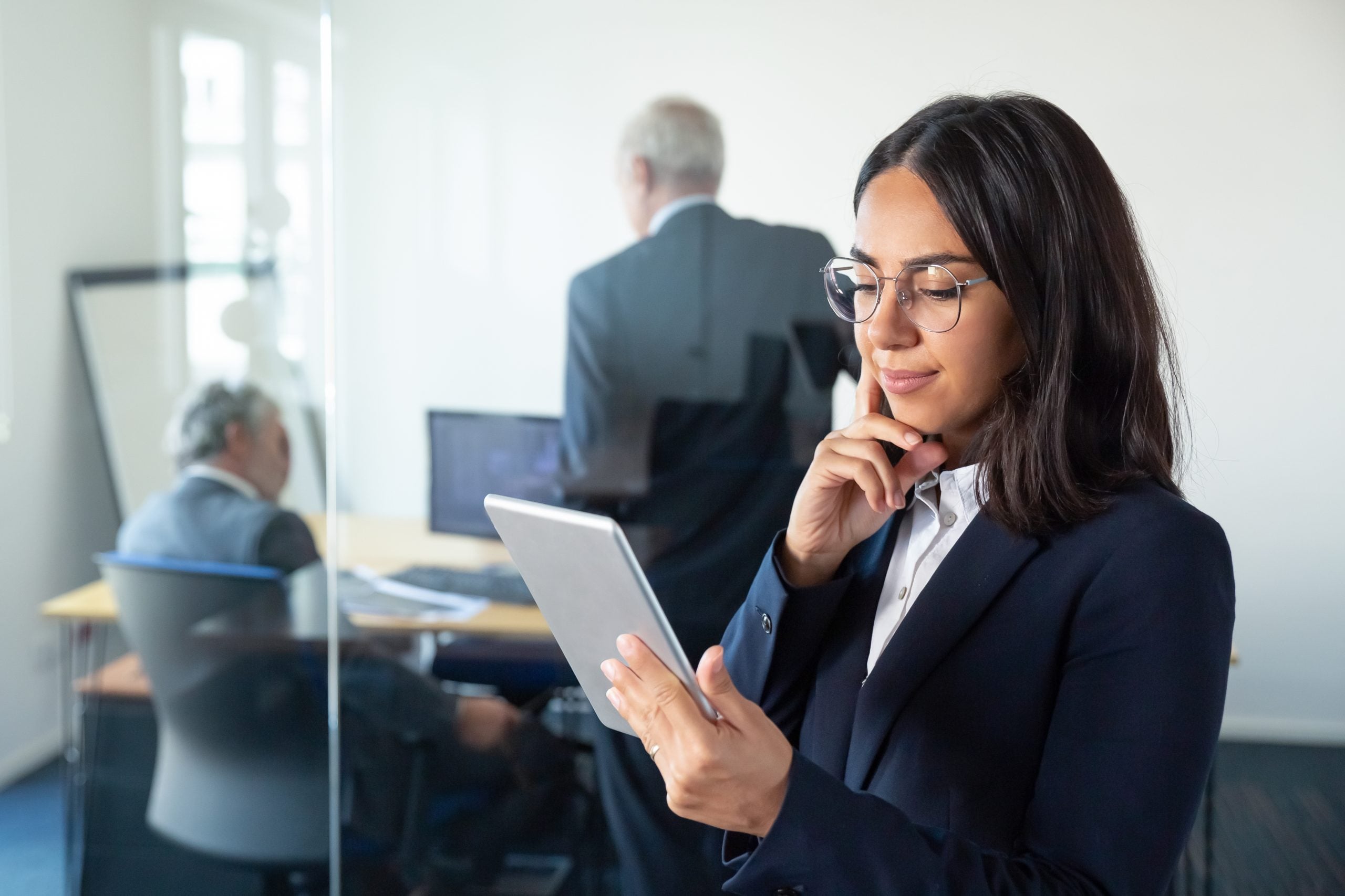 A manager in glasses studies a tablet in a busy office.