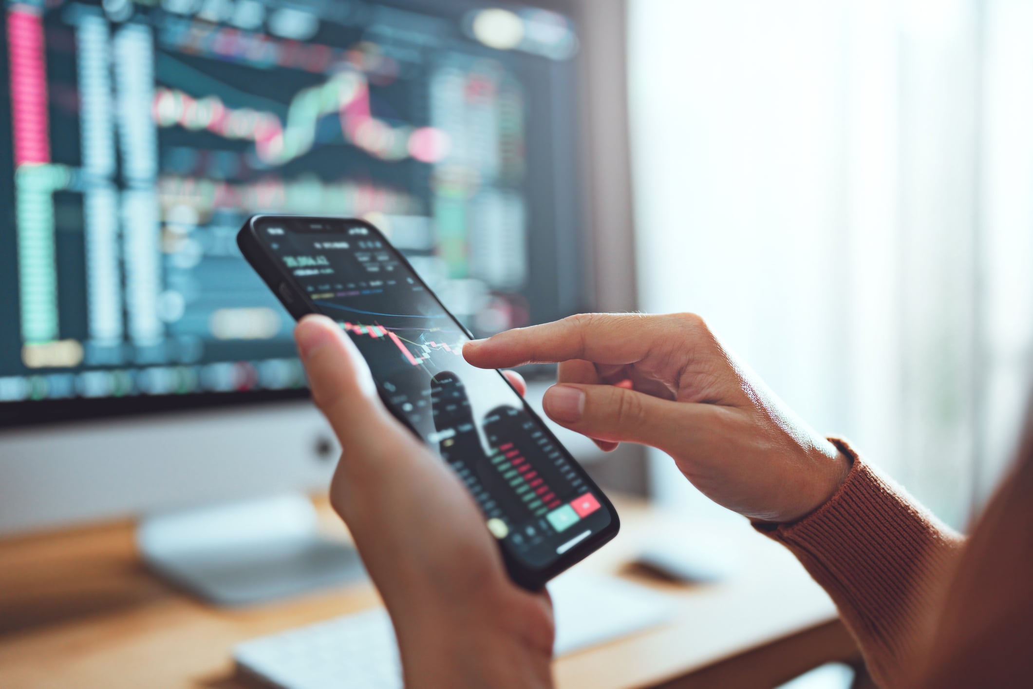 Hands checking a Bitcoin price chart on a phone in front of trading screens.